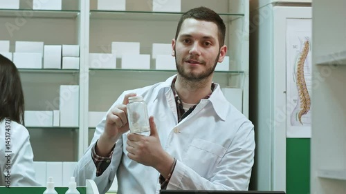 Young malepharmacist holding a white blank bottle of pills, promoting medicine, while his colleague working photo