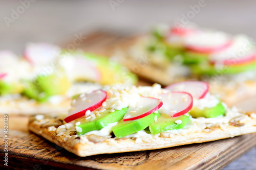 Diet and quick veggie cracker with sauce and vegetables. Fresh avocado and radishes on crispy cracker with seeds on wooden board. Closeup 