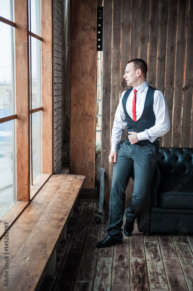 Portrait of a man in a white shirt, red tie and black vest on the background of a brick wall and a large window. Portrait of a man who is lost in thought, waiting and camera at the camera Portrait of a man in a white shirt, red tie and black vest on the background of a brick wall and a large window. Portrait of a man who is lost in thought, waiting and camera at the camera