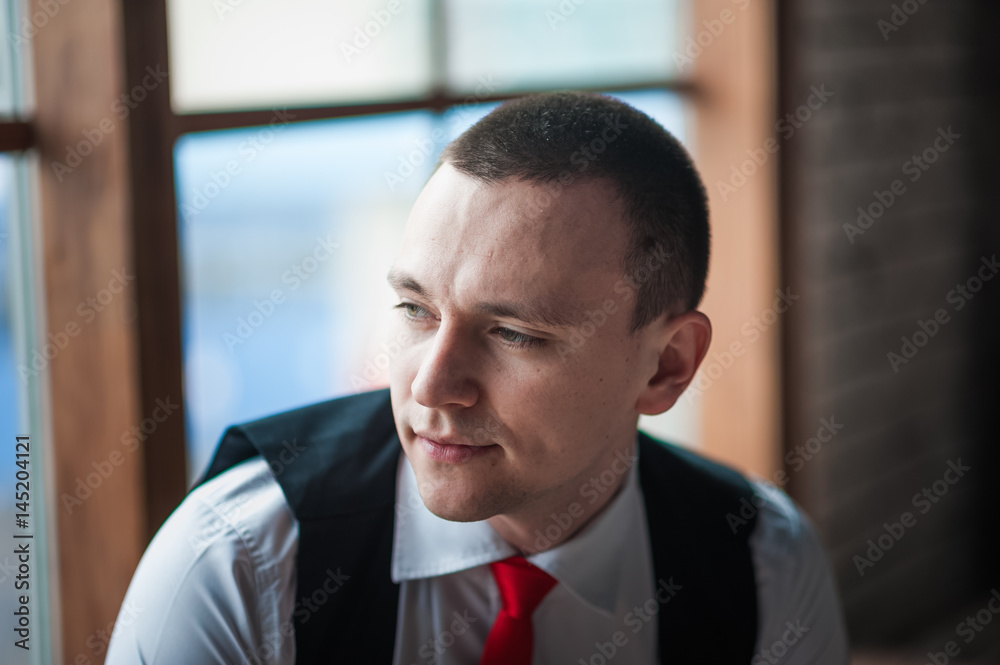 Portrait of a man in a white shirt, red tie and black vest on the background of a brick wall and a large window. Portrait of a man who is lost in thought, waiting and camera at the camera