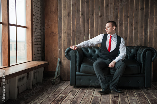 Portrait of a man in a white shirt, red tie and black vest on the background of a brick wall and a large window. Portrait of a man who is lost in thought, waiting and camera at the camera © Nelly