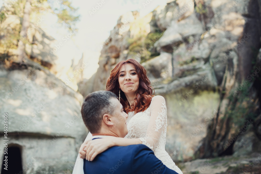 Groom in blue suit raises bride up standing on the rocks