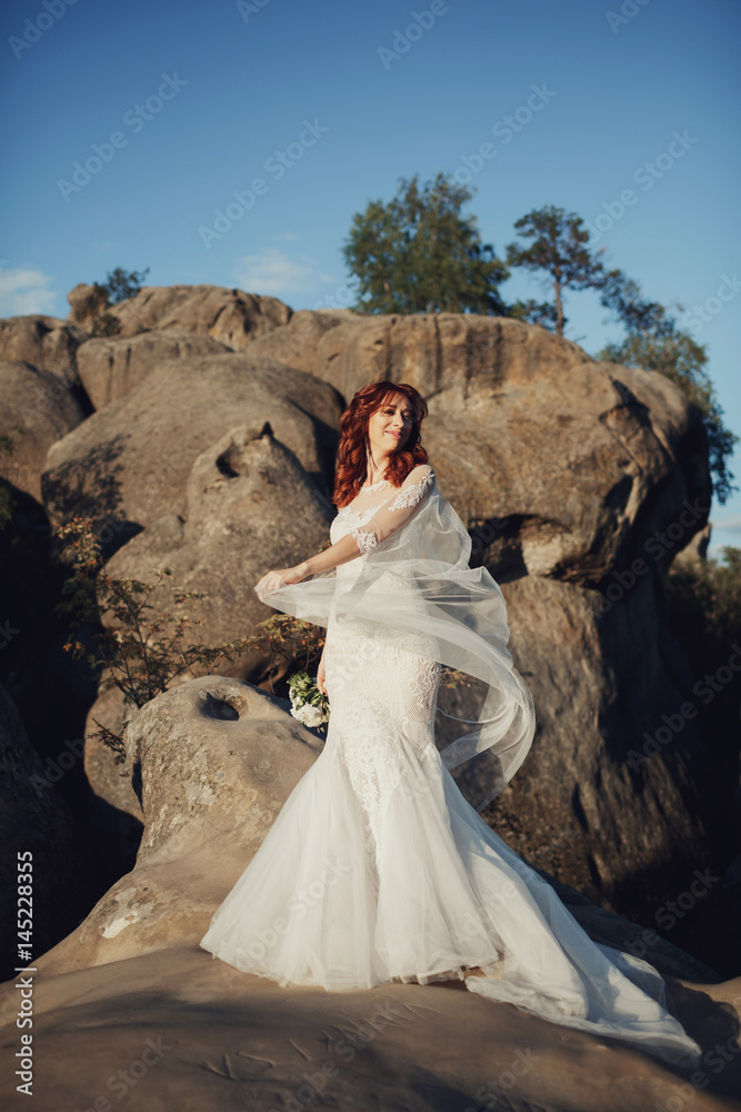 Bride with red hair poses on the rocks in rays of evening sun