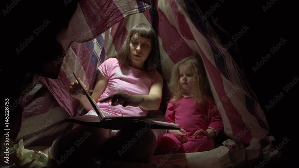 Mother and daughter in pink pajamas sit in a striped tent on the floor in a dark room at night. Tired woman reads the book to a joyful little girl in lodge before with flashlights going to bed. Mother and daughter in pink pajamas sit in a striped tent on the floor in a dark room at night. Tired woman reads the book to a joyful little girl in lodge before with flashlights going to bed.