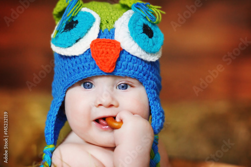 A small boy, dressed in a homemade knitted cap in the shape of an owl, gnaws a bagel