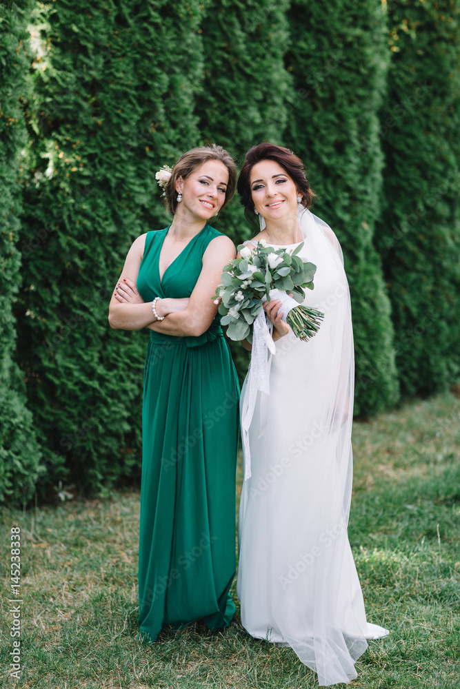 Bride and bridesmaid in green dress stand side by side on green lawn
