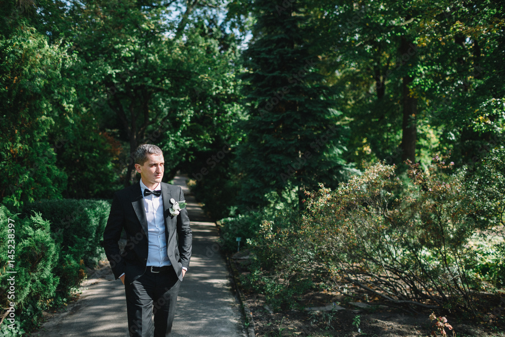 Groom in black suit with white boutonniere walks along the path in park Groom in black suit with white boutonniere walks along the path in park