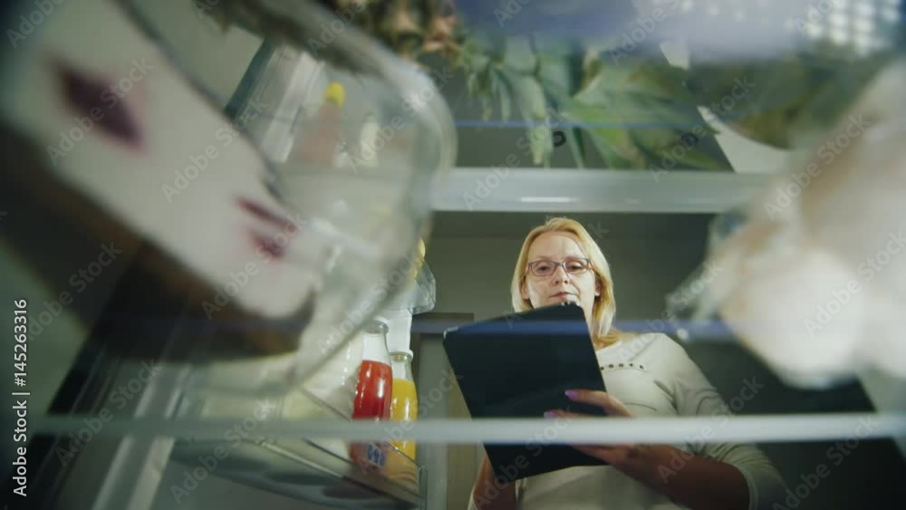 Online order food. A young woman makes a grocery list for a grocery store. He looks at food in the fridge, uses a tablet. Inside view