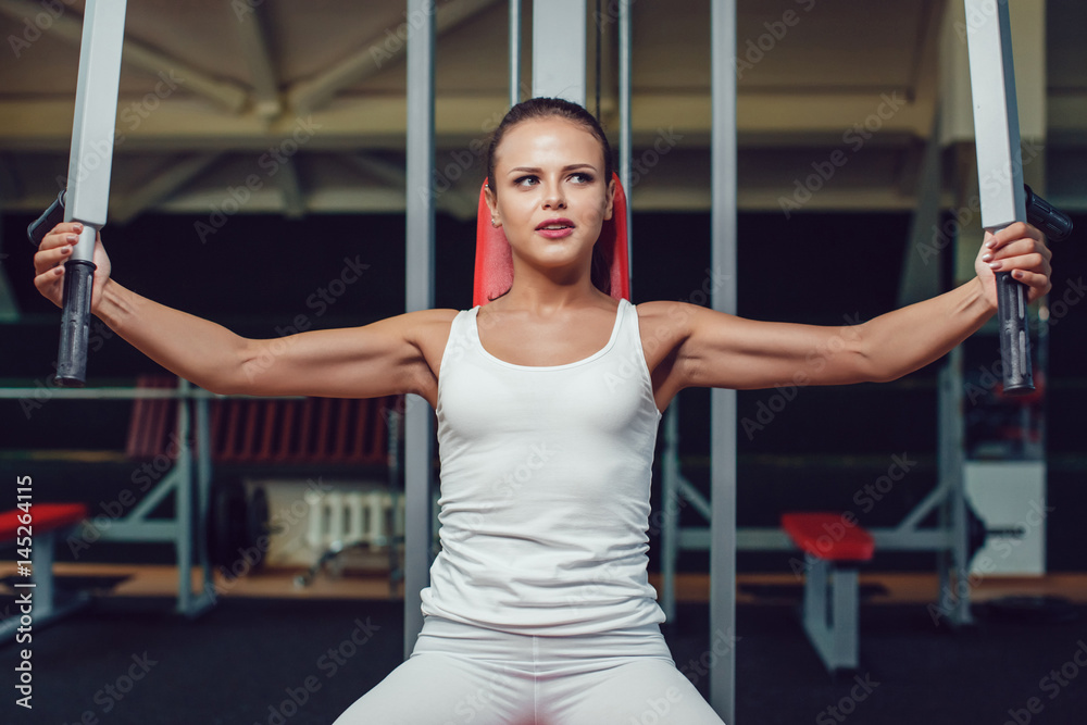Beautiful girl doing exercises on the simulator in the gym dressed blank white tank-top and tights. Beautiful girl doing exercises on the simulator in the gym dressed blank white tank-top and tights.
