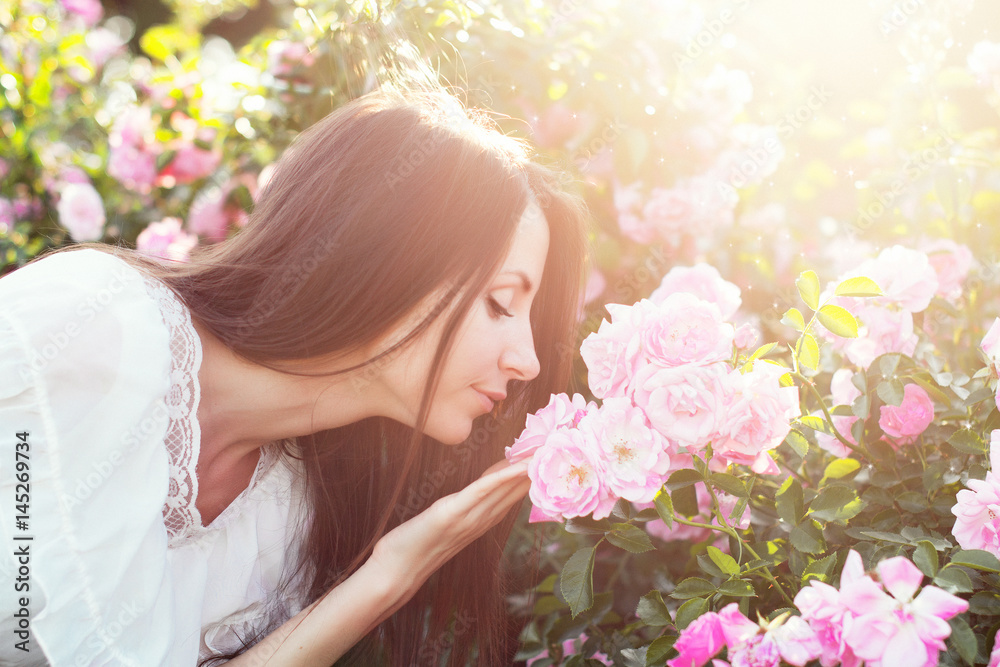 Красивая девушка с темными длинными волосами стоит возле куста роз и нюхает цветы в саду