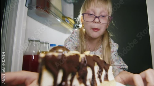 Childish pranks - a little girl in pink glasses secretly bites a chocolate cake in the fridge. Lover of sweet. Funny video
