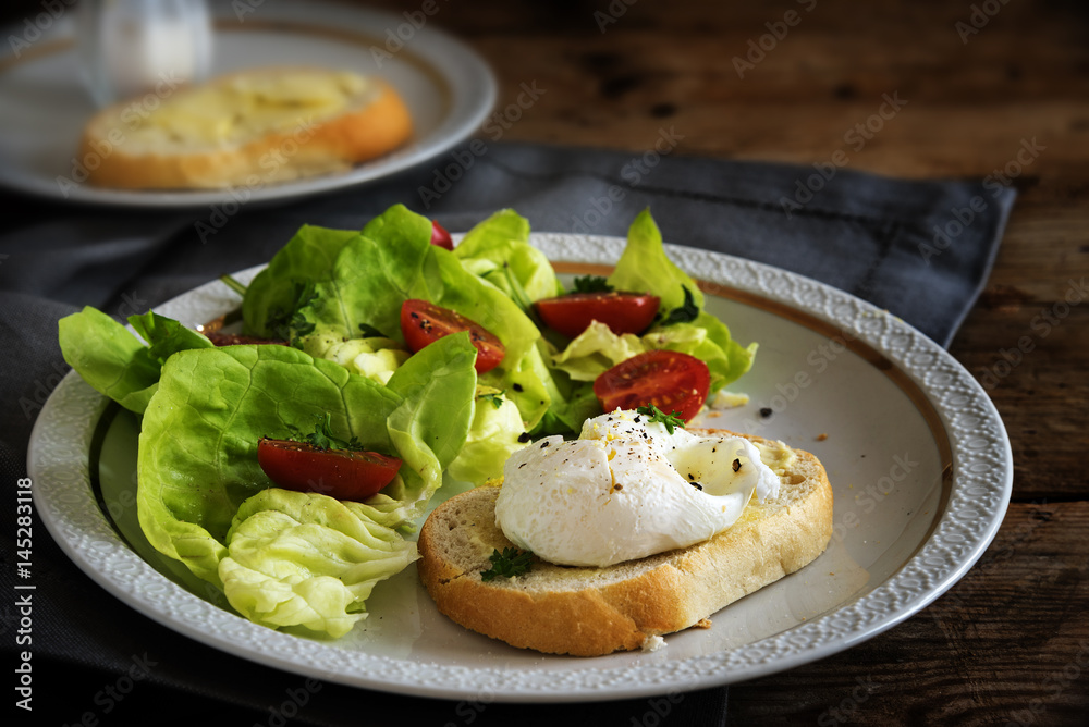 Poached egg on baguette slice with salad and tomatoes, white plate on a dark rustic wooden table, delicious breakfast or snack Poached egg on baguette slice with salad and tomatoes, white plate on a dark rustic wooden table, delicious breakfast or snack