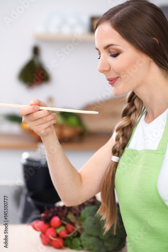 Young woman is cooking in a kitchen. Housewife is tasting the soup by wooden spoon.