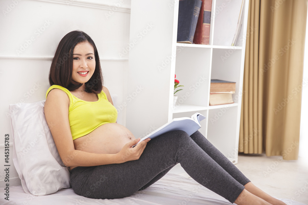 happy pregnant woman sitting on bed while reading a magazine happy pregnant woman sitting on bed while reading a magazine