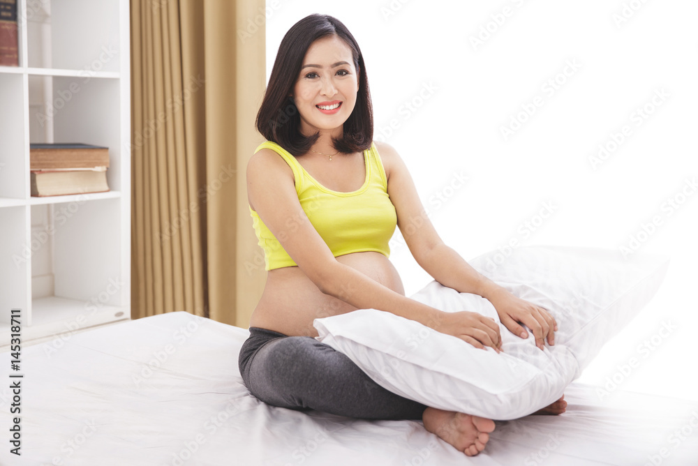 asian pregnant woman smiling while sitting on bed asian pregnant woman smiling while sitting on bed