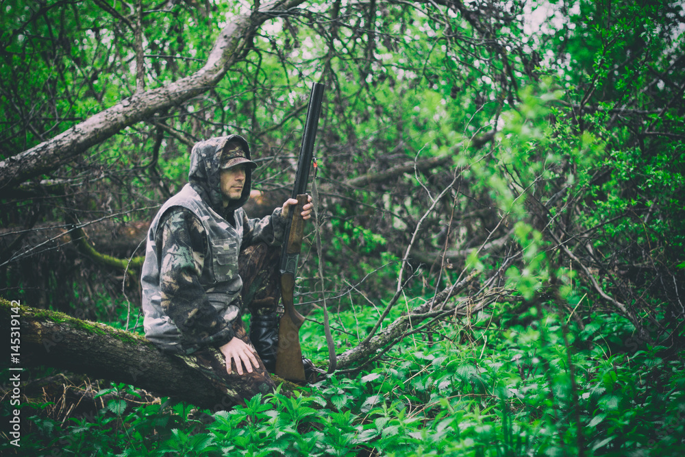 A man hunter with a gun sits on a fallen tree in rainy weather in a spring forest, toned photo
