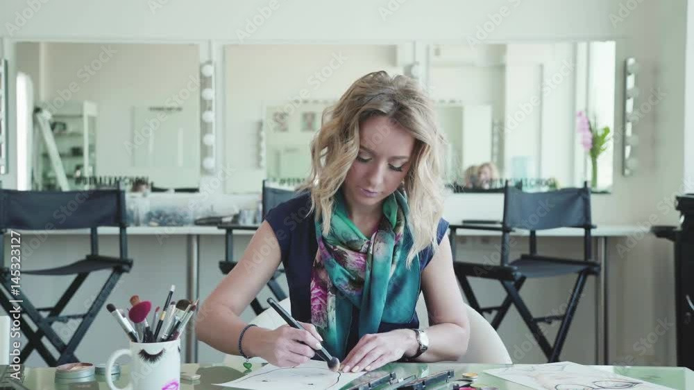 Make-up artist creates a make-up sketch on the face chart. Woman in a draws a facechart. Stylish blonde woman in a bright studio sits at a table and draws a face chart using makeup and brushes Make-up artist creates a make-up sketch on the face chart. Woman in a draws a facechart. Stylish blonde woman in a bright studio sits at a table and draws a face chart using makeup and brushes