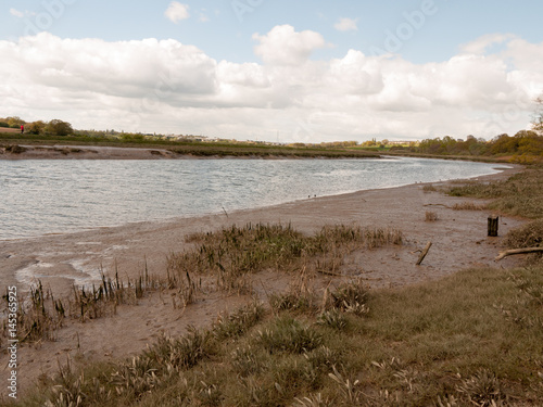 A Waterway River Through the UK with Low Tide and Birds Wading in the Water and the Mud Bank with A Cloudy Blue Sky and Brown Grassy Land