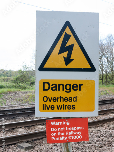 A Railway Safety Sign Saying Danger Overhead Live Wire Warning Do Not Trespass On the Railway Penalty £1000 in Black, Yellow, Red, and white with Track in Background in UK
