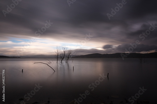 Long exposure photo of some trees and branch in still water  beneath moving clouds. A distant portion of the sky is blue  while gray dominate the whole image