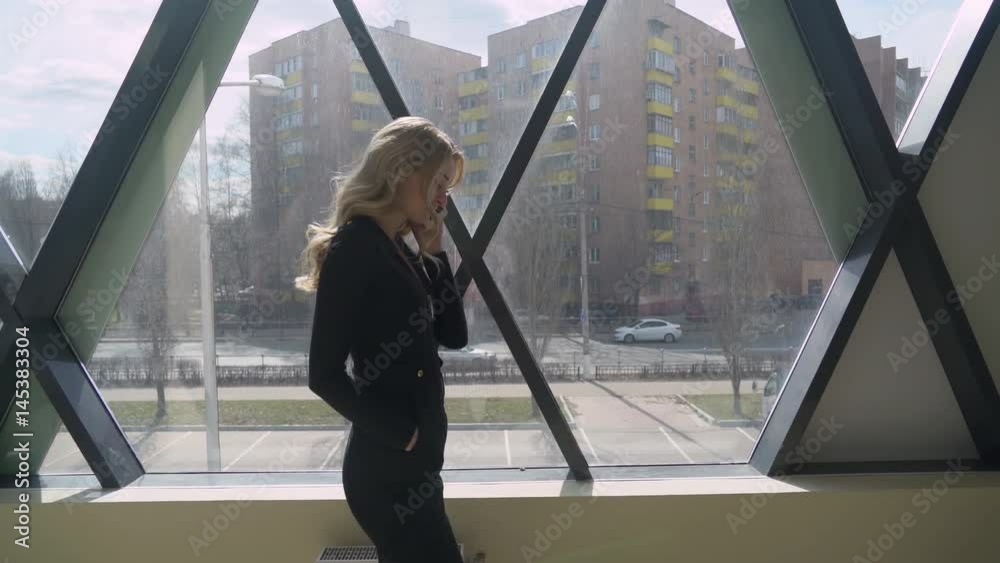 Beautisul blonde girl in black walking by the window and talking on the phone Beautisul blonde girl in black walking by the window and talking on the phone