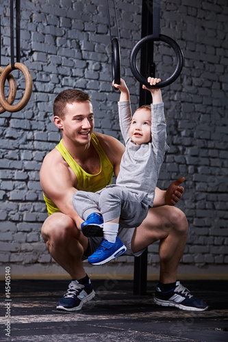 Happy father and little son exercising with gimnastic rings and smiling against brick wall in the gym.