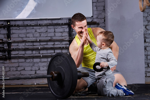 Portrait of happy father and son with barbell in the cross fit gym against brick wall.