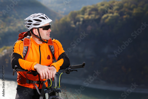 Close-up photo of cyclist in orange jacketr stands with his bike under river against beautiful landscape with mountain.
