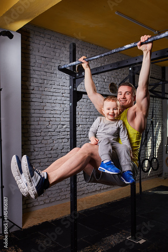 Young happy father doing pull ups on the bar with son on his legs at the cross fit gym against brick wall.