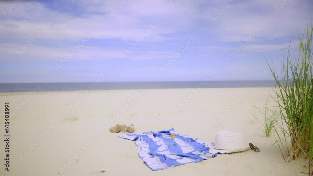 Nobody. Beach blanket with woman items outdoor on sandy beach dunes. Sea water blue sky in the background Dolly sliding shot 4K ProRes HQ codec Nobody. Beach blanket with woman items outdoor on sandy beach dunes. Sea water blue sky in the background Dolly sliding shot 4K ProRes HQ codec