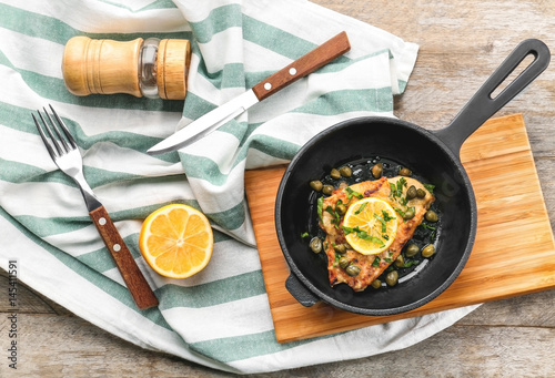 Frying pan with delicious chicken piccata on wooden board