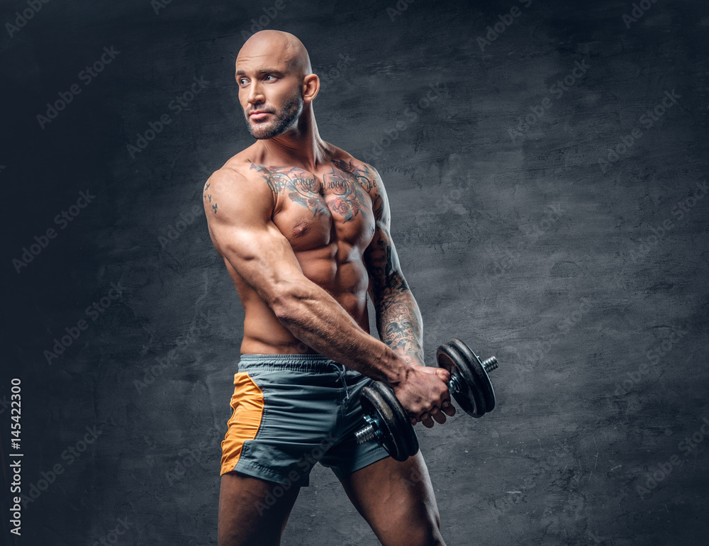 Studio portrait of athletic shirtless shaved head tattooed male dressed in a sports shorts holds dumbbell.