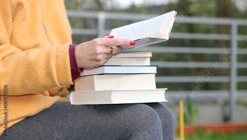 Kobieta w żółtej kurtce i szarych getrach siedzi w parku, na kolanach ma stos książek, w ręku trzyma opartą na nim otwarta książkę
