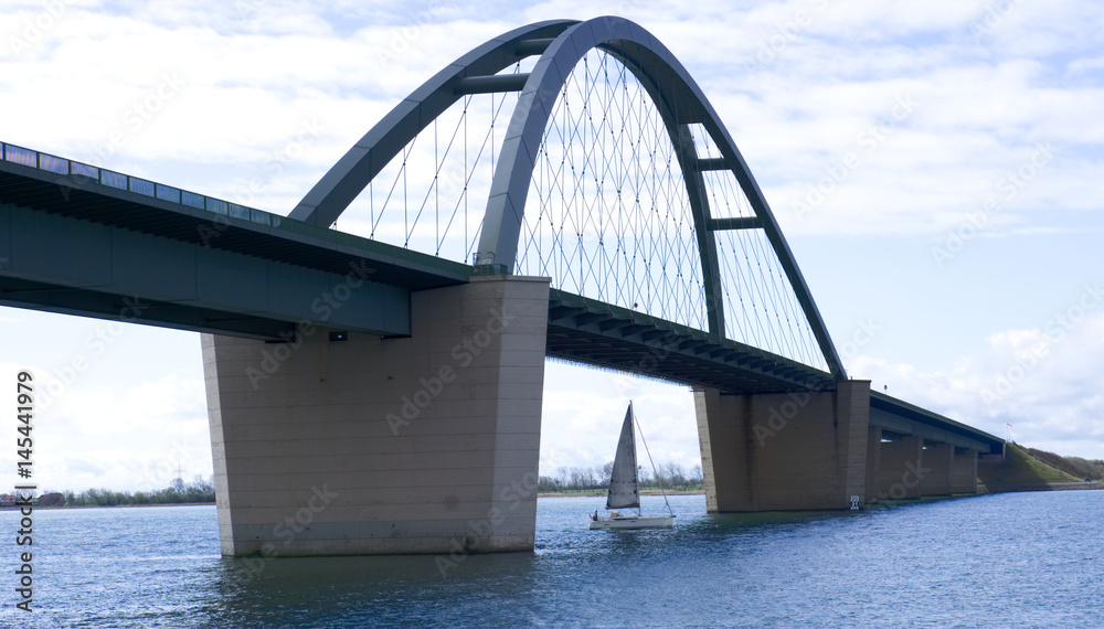 Die Fehmarnsundbrücke wird von einem Segelboot unterfahren. Eine Brücke mit Segelboot.