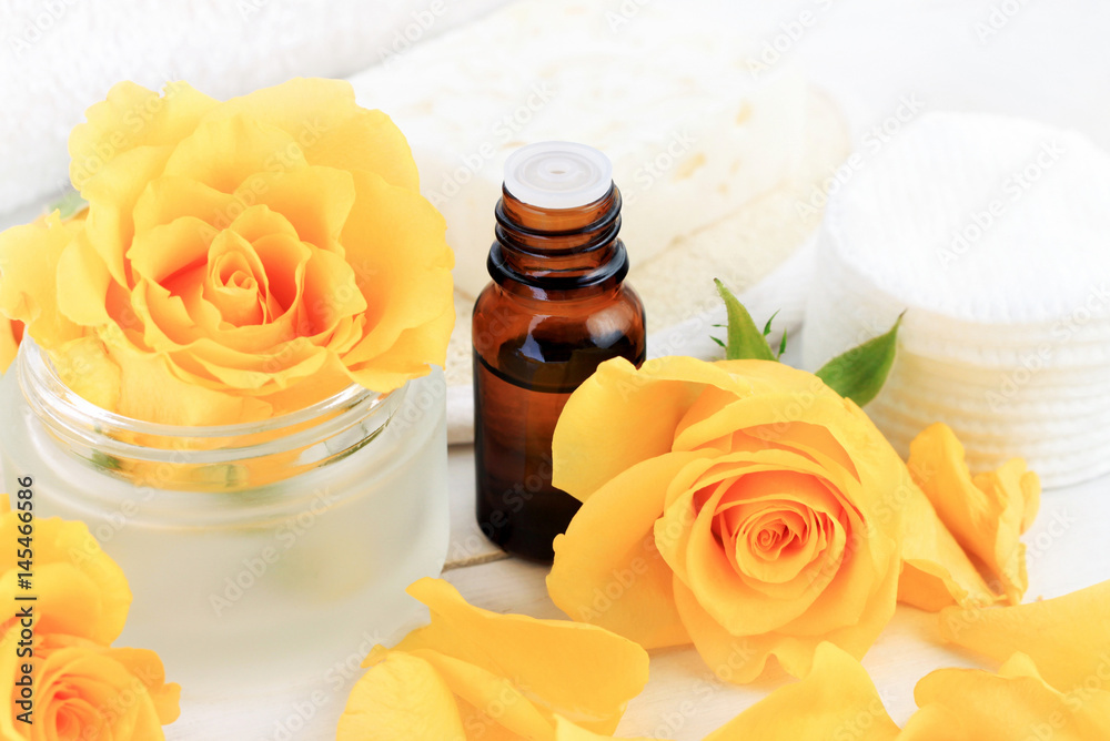 Floral aroma essence. Dark glass dropper bottle of essential oil, close up, amidst yellow roses and petals, white toiletries background. Floral aroma essence. Dark glass dropper bottle of essential oil, close up, amidst yellow roses and petals, white toiletries background.
