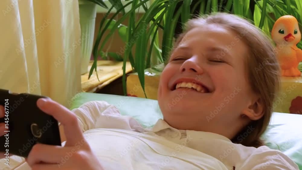 Little girl watches video on smartphone and laughs lying under cover on