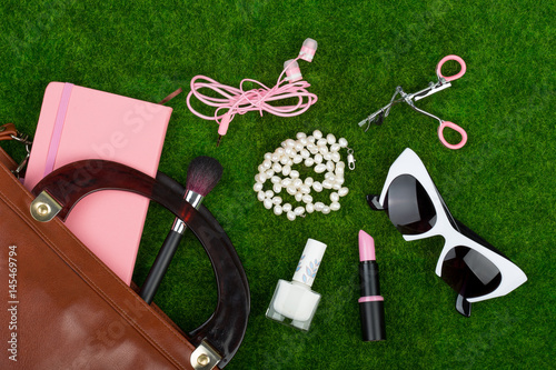 Female fashion accessories - bag, note pad, sunglasses, headphones, lipstick and other essentials on the grass