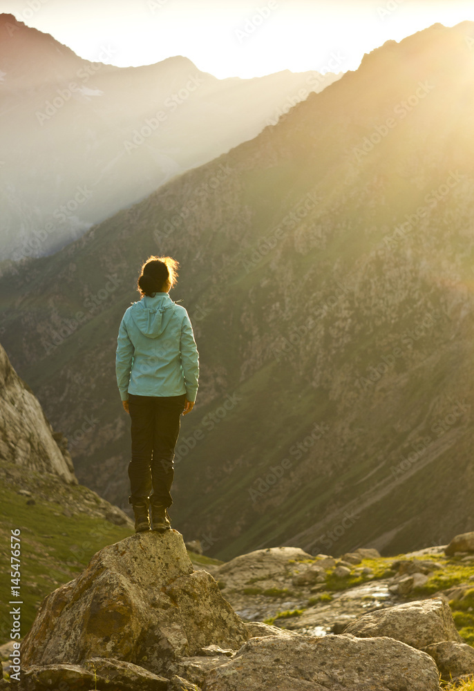 girl in blue jacket standing on stone with hands-up above mountais at sunset