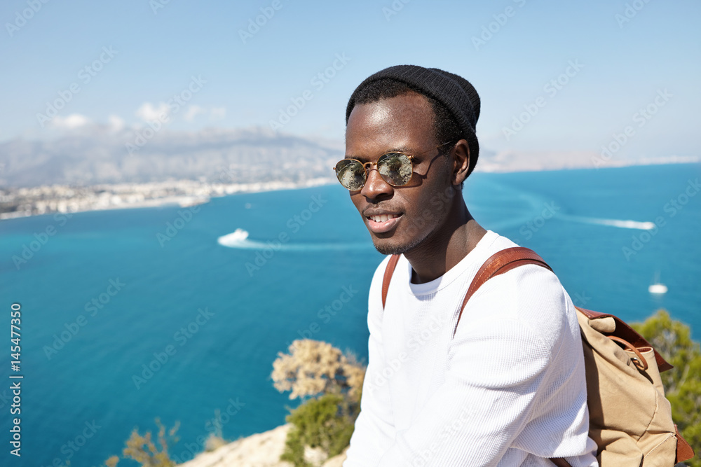 People, travel, tourism and lifestyle. Stylish dark-skinned traveler wearing trendy clothing standing on mountain above sea, enjoying beautiful amazing view, blue water, city and sky in background People, travel, tourism and lifestyle. Stylish dark-skinned traveler wearing trendy clothing standing on mountain above sea, enjoying beautiful amazing view, blue water, city and sky in background