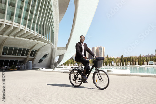 Urban lifestyle, ecology and transportation concept. Fashionable modern ecologically friendly young Afro American businessman wearing trendy round shades and formal suit cycling to work on bicycle