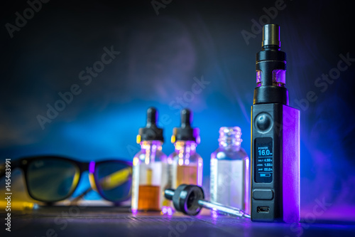 Vaping mod e-cig with tank atomizer and juice bottles over wood background photo