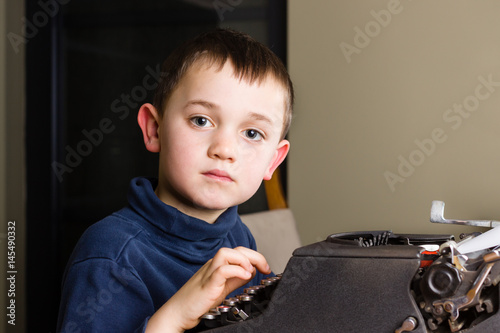 Cute little boy typing a letter on a vintage black typewriter at home, side view