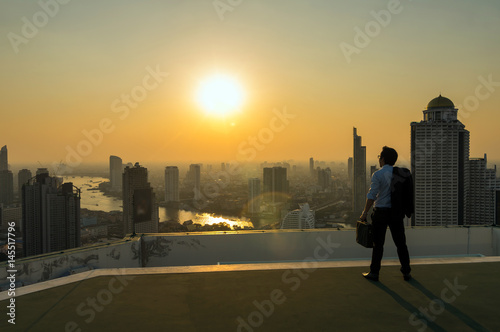 Silhouette of businessman standing with carrying the business bag and looking the vision over the cityscape background at sunset time with lens flare,Business success concept