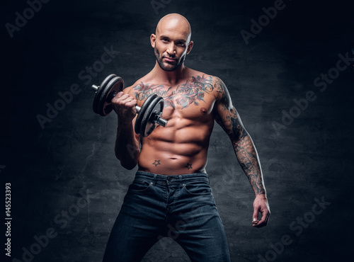 Shirtless shaved head, muscular male with tattoos on his chest and arms holds dumbbell.