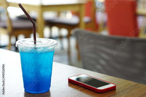 Closeup Cocktail or Tropical Mocktail - Plastic glass of Blue Hawaii Italian Soda with lemon and moblie phone on wooden table and blurred background.