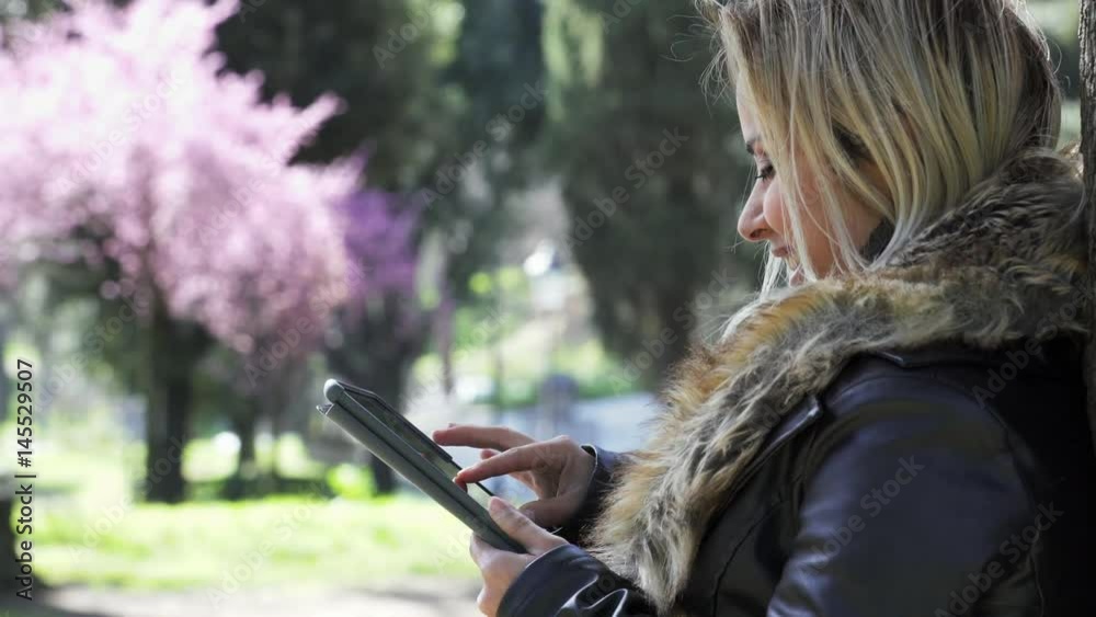 Smiling woman profile leaning against a tree using tablet, park in background Smiling woman profile leaning against a tree using tablet, park in background