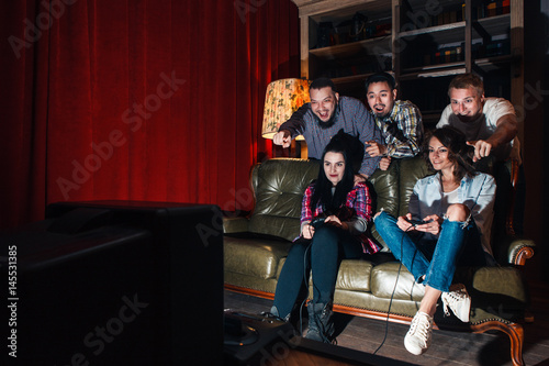 Group of youth have fun playing video game, competition at home. Two girls with joysticks on couch, three guys cheer emotionally, free space