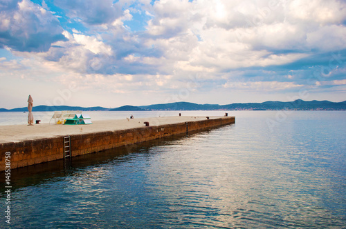 A stone pier with metal ladders in the rippled sea water against a hill range in the distance and dramatic cloudy morning sky. Zadar  Croatia