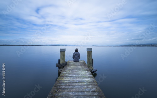 Overcast. Self reflection in magical world of fantasy. One woman sits on a wooden pier. Cloudy above the lake. Long exposure.
