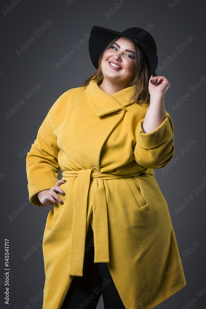 Fototapeta premium Plus size fashion model in yellow coat and black hat, fat woman on gray background, overweight female body